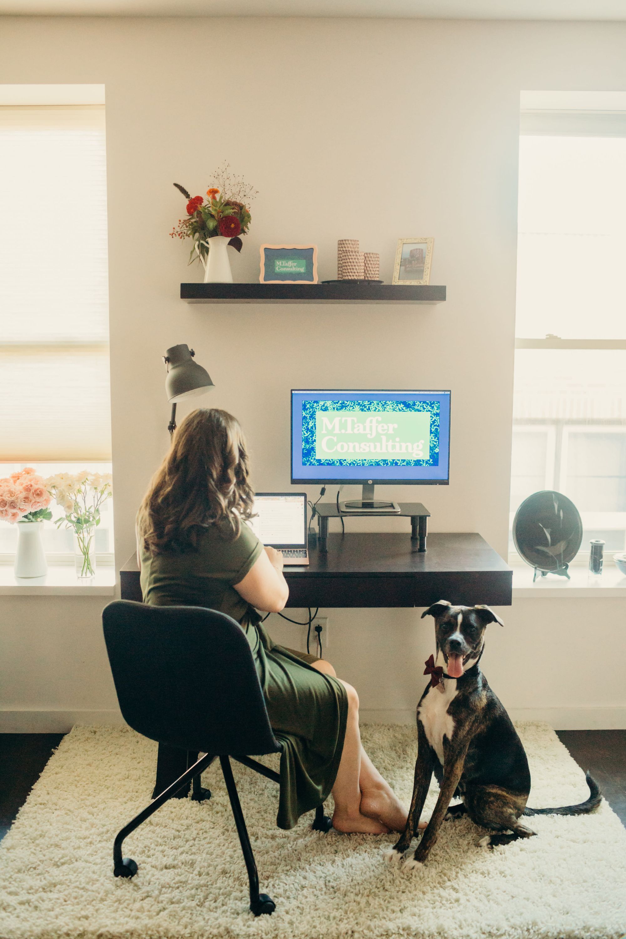 A girl sits at her desk typing with her dog at her feet. 