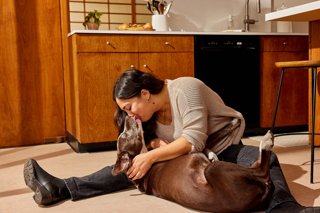 A woman with black hair in a gray sweater sitting on the floor of a mid century modern kitchen and kissing her senior dog who has a sleek, brown coat.