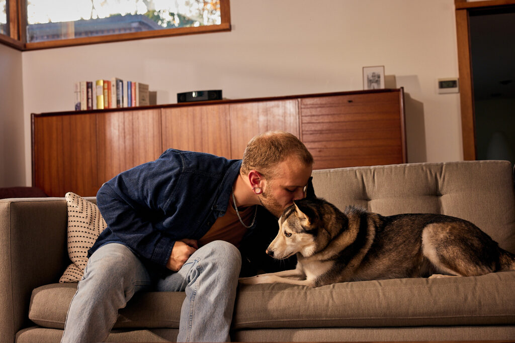 A man in a blue shirt and jeans leaning over to kiss his husky dog on the forehead. They are sitting on a gray couch in a mid-century modern living room.