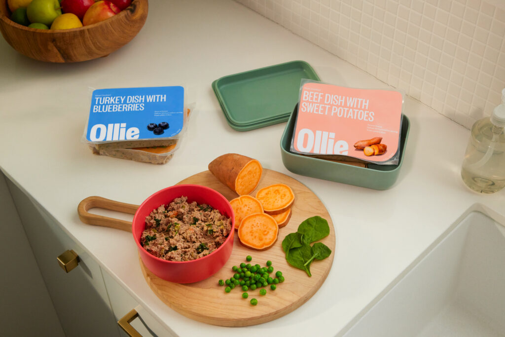 A bowl of Ollie fresh dog food arranged on a wooden cutting board with sweet potatoes and spinach. Nearby are Ollie fresh dog food packs on a white countertop.