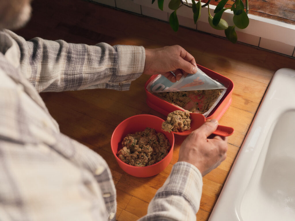 A man preparing Ollie fresh dog food by scooping it from the package into a bowl while standing at a wooden countertop