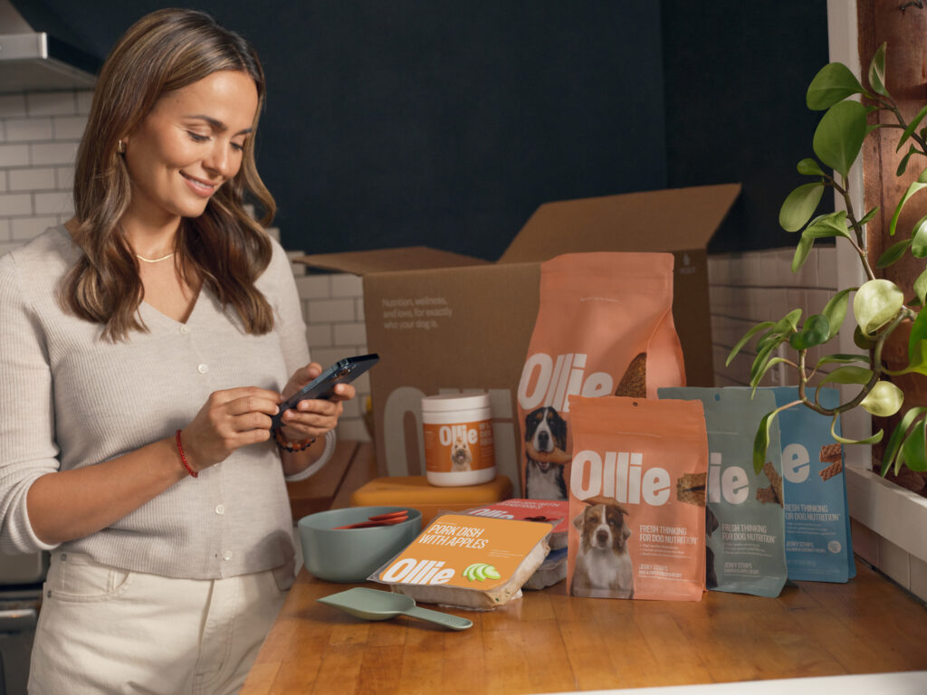 A brunette woman in a cream sweater reading her phone in a modern kitchen as she stands next to a box of Ollie products including Ollie fresh dog food, Ollie baked dog food, Ollie treats and supplements.