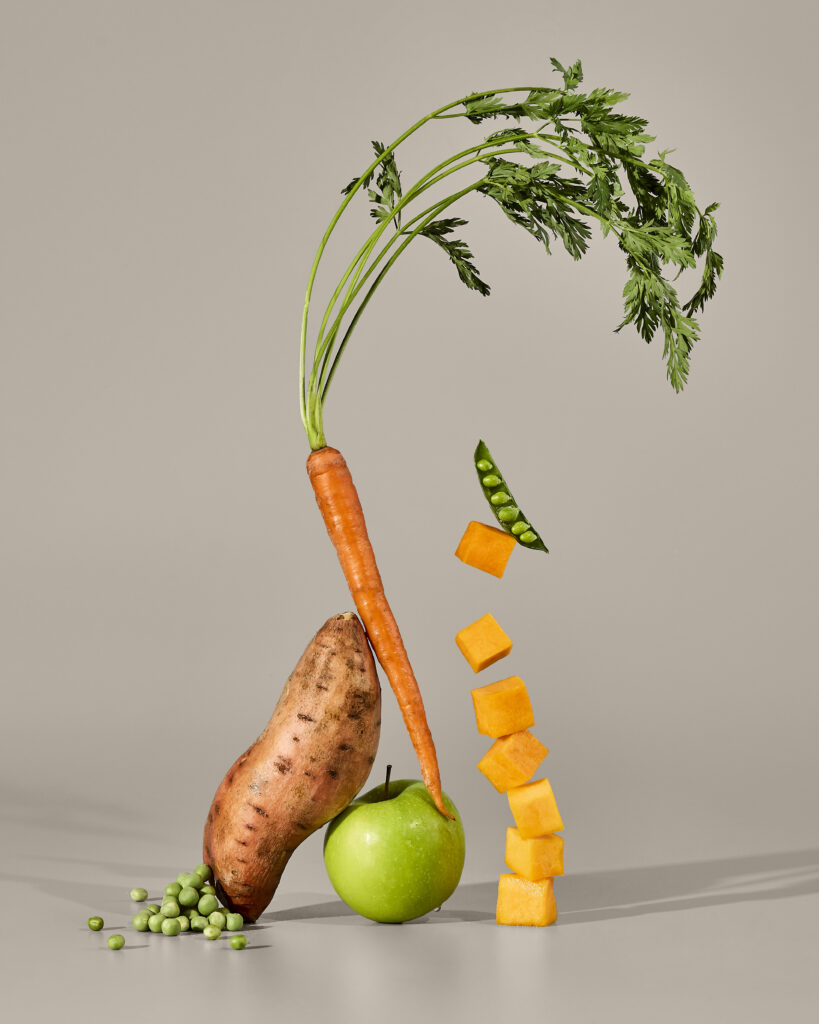 A still life of balancing sweet potato, carrot, peas, green apple, and cubed butternut squash on a gray seamless background.