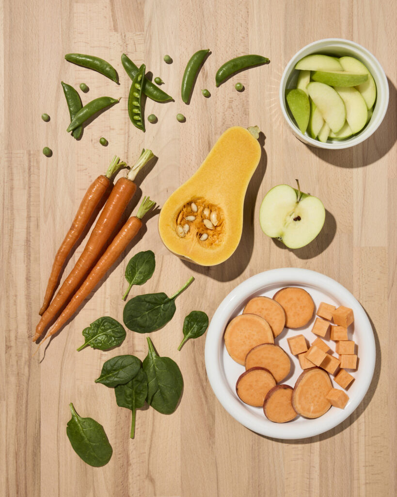 An overhead photo of ingredients displayed on a wooden countertop. Fresh carrots, peas, spinach, butternut squash, sweet potato, and green apples.