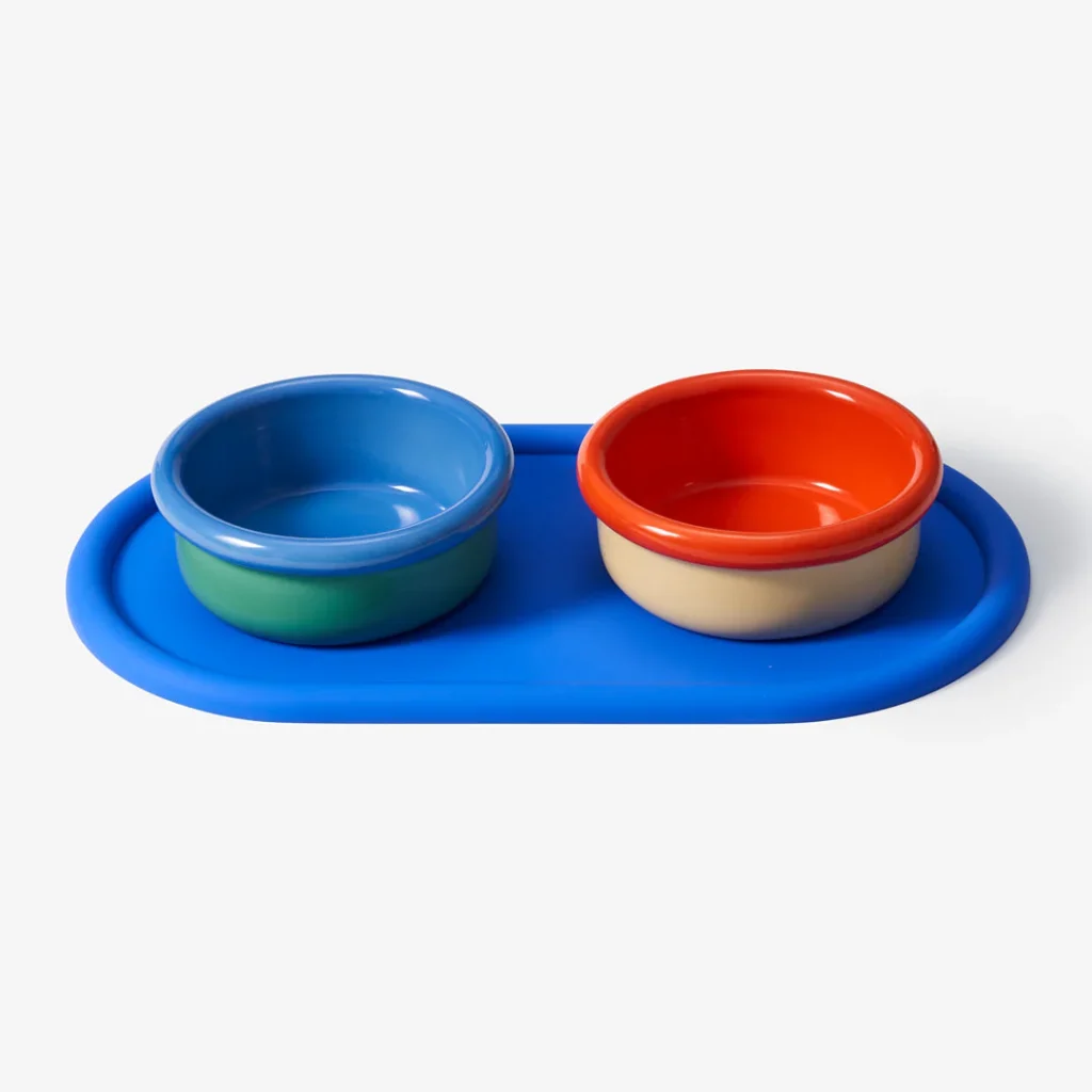 A bright blue rounded tray, holding a blue and green ceramic dog bowl on the left, and a red and cream ceramic dog bowl on the right.