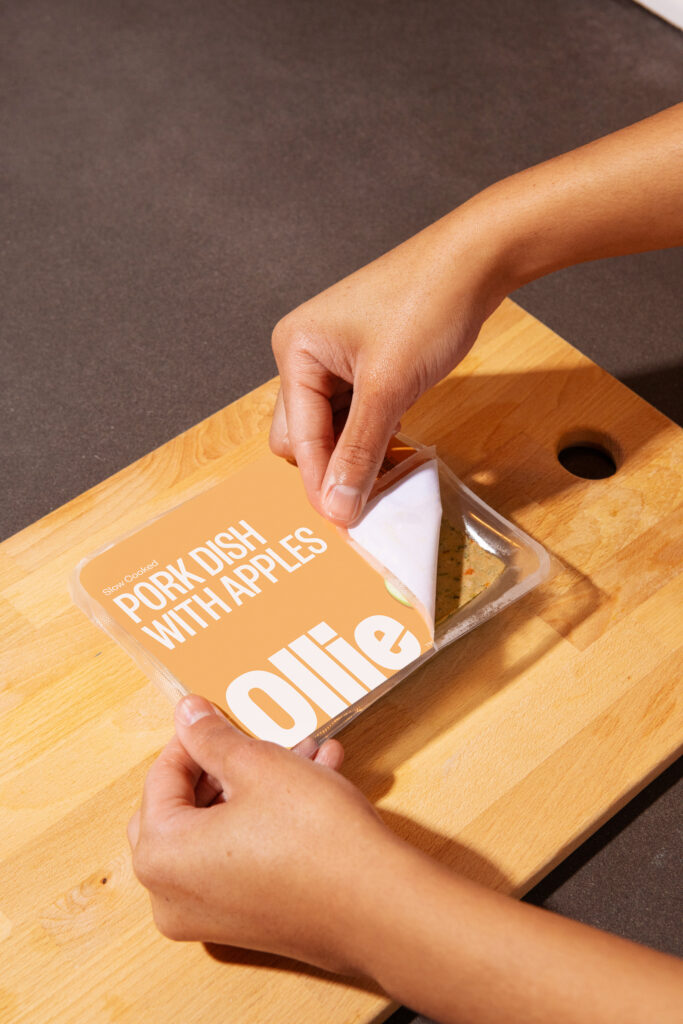 A pack of Ollie fresh dog food, pork dish with apples, on a wooden cutting board, being peeled open by a person