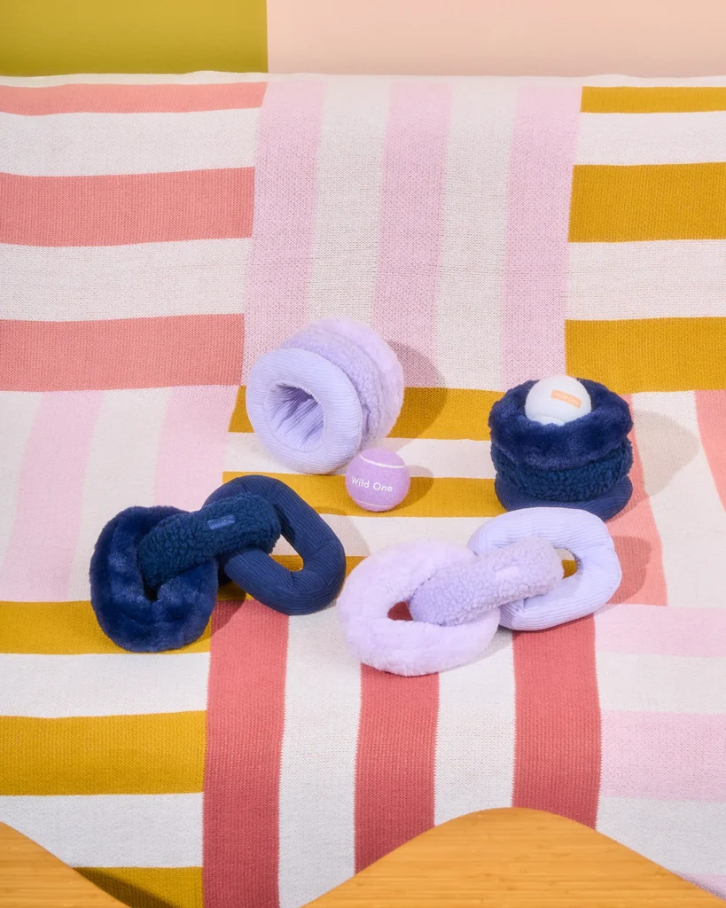 Four plush dog toys shaped as chain links and coils, colored lavender and navy, photographed on a multi-colored and striped blanket.