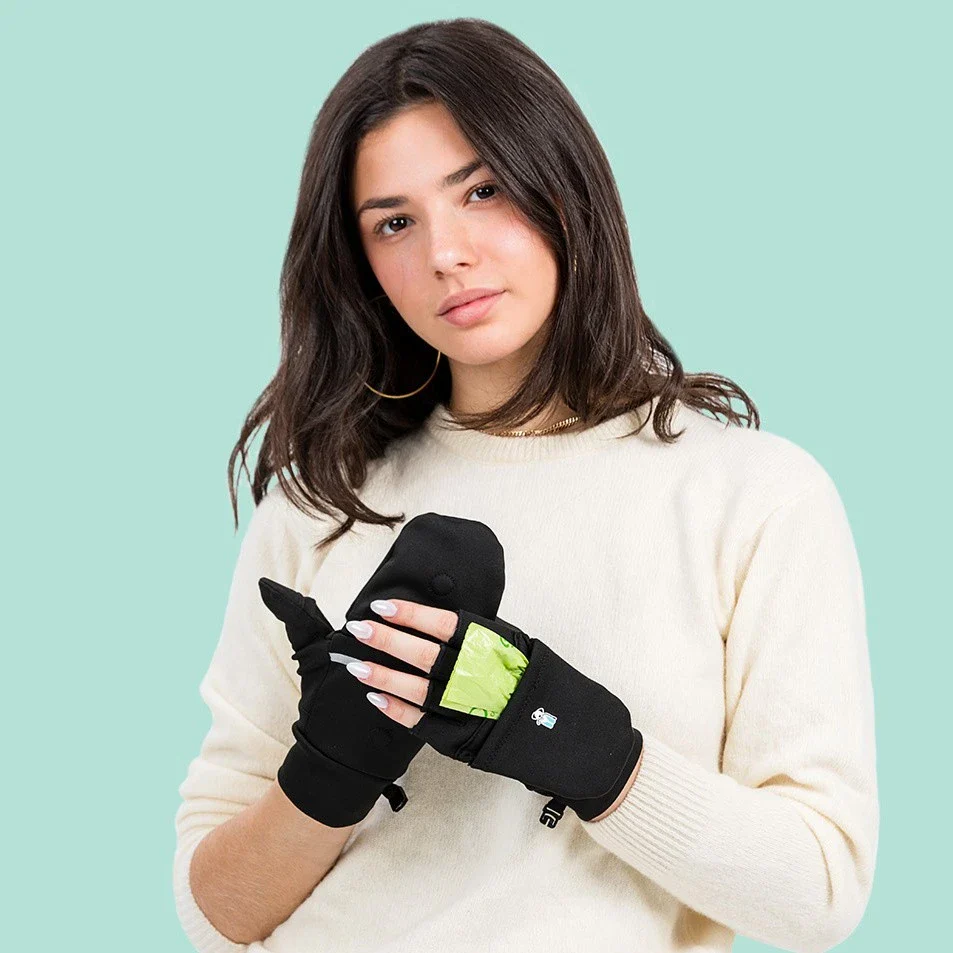 A female model wearing a pair of black Walkee Paws mitten gloves. Her  left hand's fingers are exposed, showing the gloves' interior pocket for dog poop bags.